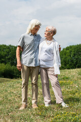 full length of happy senior husband and wife with grey hair standing and hugging on green hill in summer.