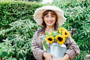 Portrait of smiling young female farmer woman holding watering can with fresh sunflowes bouquet on the green trees background. Cottagecore lifestyle. Selective focus, copy space.
