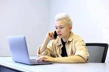 Girl with short blonde hair sitting at laptop and talking on mobile phone. Tomboy lifestyle,...