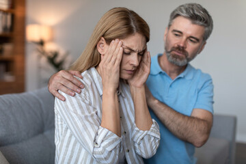 Sad adult caucasian man with beard calm offended wife, lady suffers from migraine in living room interior