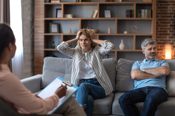 Obraz premium Angry middle aged caucasian wife holding her head, freaking out, husband ignores lady at meeting with psychologist