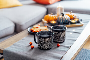 Cozy halloween plans at home. Hot tea drink in black mugs with spider net pattern, cookies and and sweets on the plate, pumpkin decor on the coffee table. Festive autumn, fall mood home decor