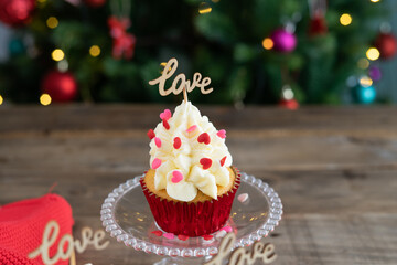 Decorated cupcake in display stand on wooden background with LOVE sign and out-of-focus Christmas tree in the background. Copy space.
