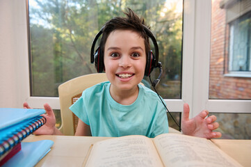 Close-up of a cheerful intelligent student of a primary online school, in headphones, smiles at camera while doing literature homework. Open to new knowledge in new semester of the new academic year