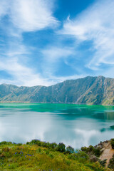 Vertical photo of the Quilotoa lagoon crater in Latacunga, Ecuador. Turquoisewater reflecting the blue sky during a sunny day. Freedom and tranquility concept.
