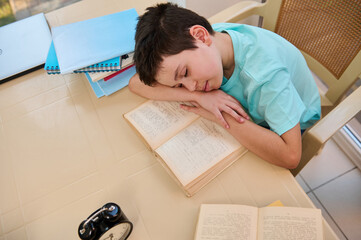 Top view of a tired sleepy schoolboy falling asleep on a textbook while doing homework. Homeschooling. Daily routine of a student. Learning disability. Difficulties in restoring the school regime