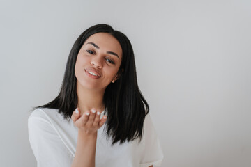 Adorable African American woman in white t-shirt sending air kiss, looking at camera, smiling on grey background with empty space. Satisfied Brazilian girl isolated. Mockup beautiful people in love.