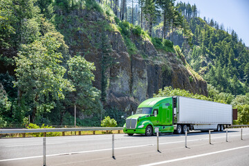 Bonnet bright green big rig semi truck with high roof cab transporting cargo in reefer semi trailer driving on the awesome highway road along the mountain with forest in Columbia Gorge