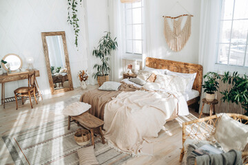 Cozy rustic bedroom with boho ethnic decor. Enter apartment with sun rays from large windows and wooden furniture. Console table, full-length mirror and handmade textiles. Plants in pots. Nobody