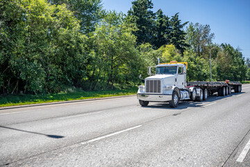 Day cab classic big rig with oversize load sign on the roof transporting heavy cargo on flat bed semi trailers driving on the wide highway road with trees on the side