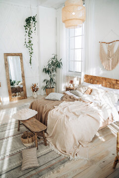 Cozy rustic bedroom with boho ethnic decor. Enter apartment with sun rays from large windows and wooden furniture. Console table, full-length mirror and handmade textiles. Plants in the interior