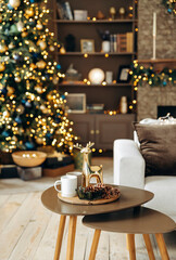 A coffee table with Christmas decor on a blurred background of a sofa and a Christmas tree with garland lights in the cozy room with a fireplace and New Year's decor. Christmas deer and fir cones.