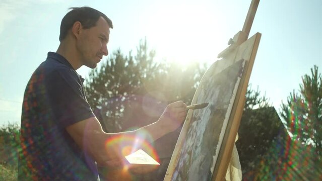 A male artist paints a picture on an easel with a brush. Sunlight and inspiration in a beautiful landscape of nature and a person who draws and relaxes. High quality 4k footage