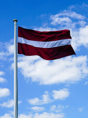 Fototapeta na wymiar Latvian National Flag on the mast waving in the wind against blue sky with white clouds