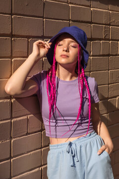 Portrait of a caucasian teenage hipster girl in a lilac panama hat with pink braids against a pink brick wall.Summer concept.Generation Z style.