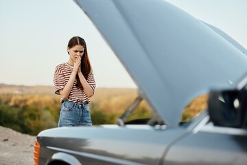 Woman traveler standing near the car with the hood open and looking for the cause of the car breakdown alone without men on the road in the countryside