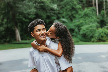 A little sister on her big teenage brother's back giving him a kiss on the cheek in an endeering...