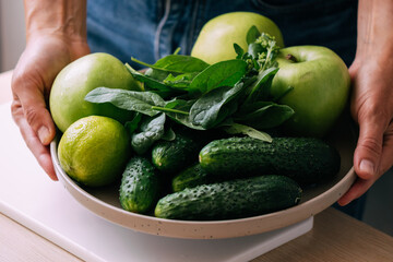 A woman holds in her hands fresh green vegetables and fruits in a large plate, close-up soft focus