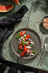 Grilled organic  watermelon salad with goat cheese and red onion, basil on textured dark table with shadows, summer fruit salad, top view, copy space.