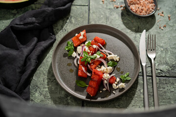Grilled organic  watermelon salad with goat cheese and red onion, basil on textured dark table with shadows, summer fruit salad, top view, copy space.