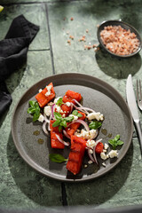 Grilled organic  watermelon salad with goat cheese and red onion, basil on textured dark table with shadows, summer fruit salad, top view, copy space.