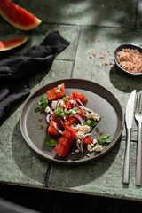 Grilled organic  watermelon salad with goat cheese and red onion, basil on textured dark table with shadows, summer fruit salad, top view, copy space.