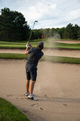 Male golfer in gray with ball in sand trap, bunker shot, multiple bunkers, end in sight, overcoming obstacles, challenging shot, target in sight, sand in air, post-swing