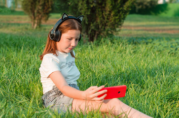 A teenage girl sits in a park on the grass with a smartphone in her hands. 12 year old girl listening to music in headphones sitting in the park on the grass
