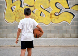 A cute young boy plays basketball on the street playground in summer. Teenager in a white t-shirt with orange basketball ball outside. Hobby, active lifestyle, sports activity for kids
