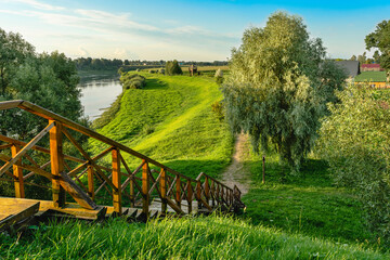 A wooden gangway of an old fortress on the river bank among lush trees and rural houses. Scenic landscape with green grass and blue sky in summer evening