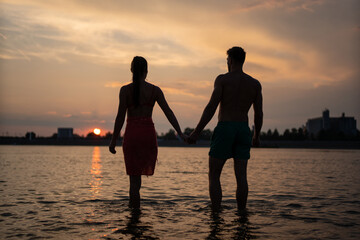 Silhouette of young couple in love enjoying the sunset on the river beach. A beautiful couple walking and relaxing on a river beach near the city after a hard day on work.
