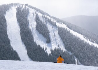 Skier in an orange jacket sitting on top of a snowy mountain and observing the beautiful view
