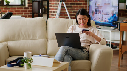 Fototapeta na wymiar Joyful woman inserting credit card credentials on on shopping site for online payment while working remotely. Asian customer sitting on couch at home while paying for products on digital store.