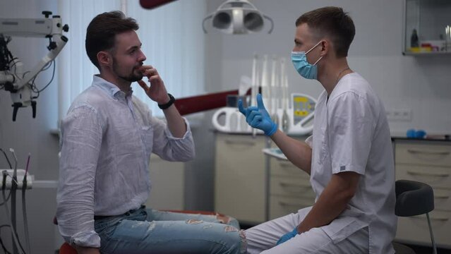 Side view dentist asking questions talking with Caucasian man sitting in dental chair. Portrait of professional confident doctor consulting patient in medical clinic indoors. Slow motion