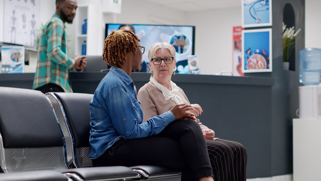 Senior patient and african american woman talking in waiting area at medical facility reception. Diverse people sitting in hospital waiting room to attend healthcare checkup visit. Handheld shot. - Powered by Adobe