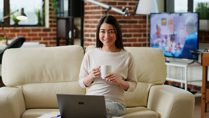 Asian woman sitting in living room working from home while smiling at camera. Happy student inside modern apartment having laptop on table while waiting for online class to start.