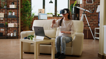 Playful woman with VR goggles playing on metaverse inside living room. Young adult person wearing futuristic virtual reality headset enjoying cyberspace while sitting on sofa at home.