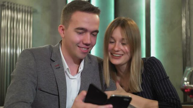 An attractive couple is sitting at a table using a smartphone, browsing the web, discussing online purchases, ordering food from a restaurant for a romantic dinner