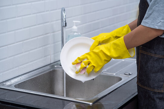 Closeup Of Woman Hands Wearing Yellow Rubber Gloves Washing Containers On Sink In Kitchen. Woman In Kitchen Rinsing White Plate In Washbasin.