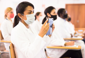 Interested hispanic female doctor in protective face mask attending refresher course, using...