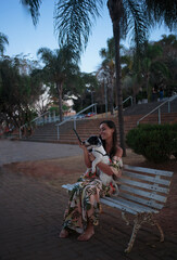 woman sitting, reading a book, on a park bench with a pet dog
