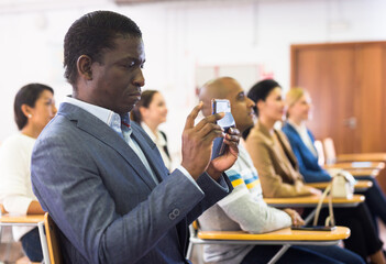 Portrait of focused man participant of business event recording lecture on smartphone at conference...