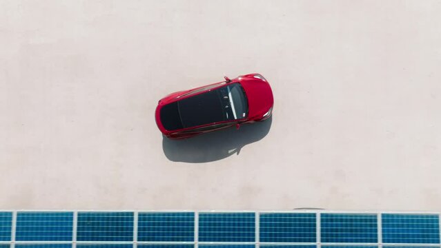 Generic red electric car driving by electric vehicle charging station under blue solar panel batteries at rooftop parking lot. Zero emission vehicle at EV only charging station on sunny summer day 4K