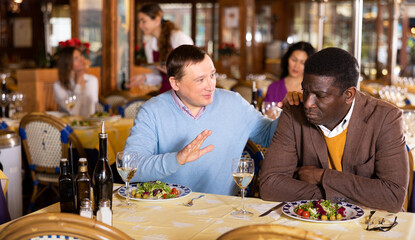Man comforting his upset African American friend during friendly meeting over dinner in restaurant..