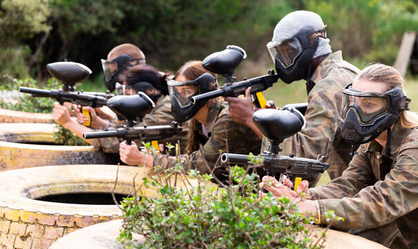 Five paintball players in camouflage and protective masks aims with guns outdoors