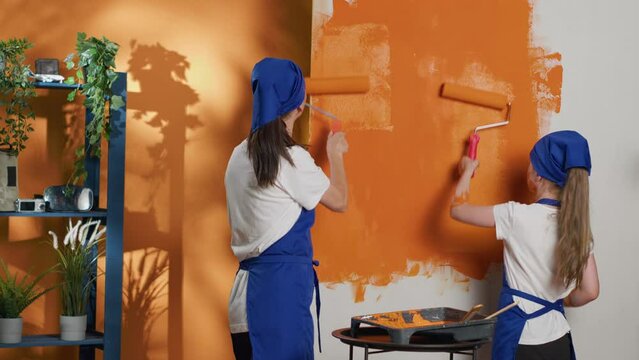 People painting walls with orange color and roller brush, using tools and equipment to paint home interior decor. Mother and young child having fun redecorating apartment room. Tripod shot.