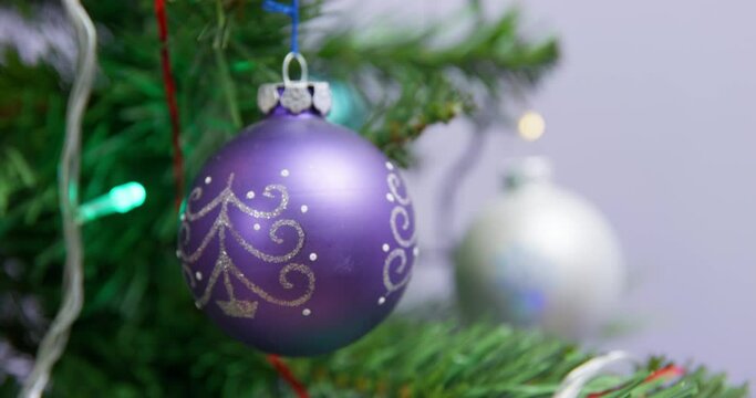 Purple Christmas ball with silver pattern hangs on evergreen fir tree near garland on blurred background in room. Festive decoration at home close view