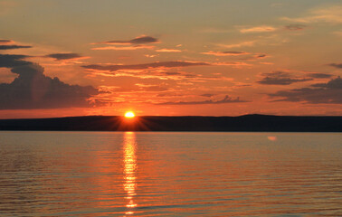 The sun setting behind the hills paints orange clouds over a picturesque reservoir.