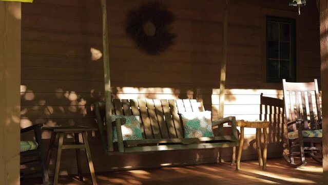 Wide shot of a wooden porch swing swaying on a beautiful summer morning in front of rural country cabin. 