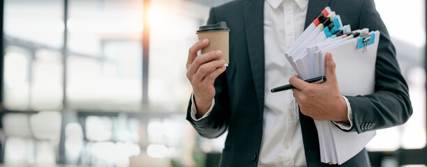 Businessman in suit holding a large stack of documents and coffee cup standing in office with copy...